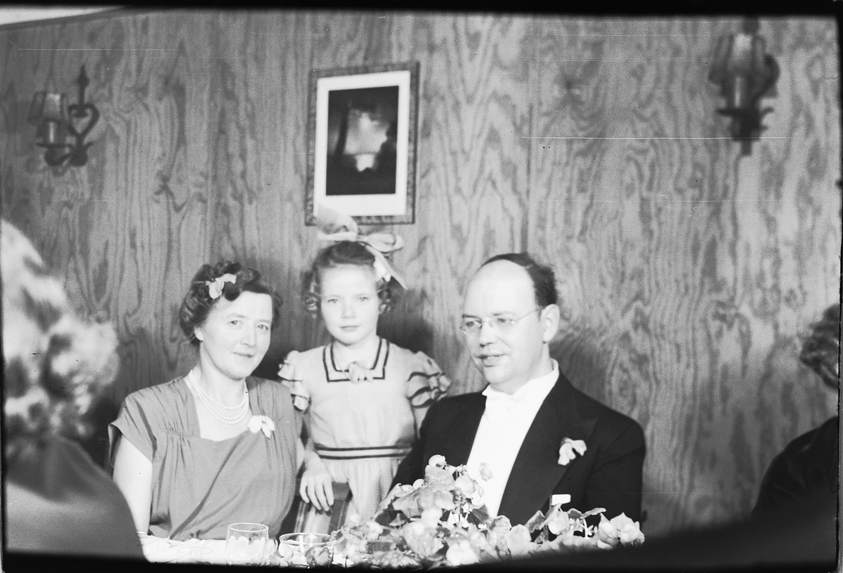 Håkon Prestkværn, Arendal, sølvbryllup. Ukjent kvinne og mann sitter til bords. Jente med sløyfe står mellom dem.