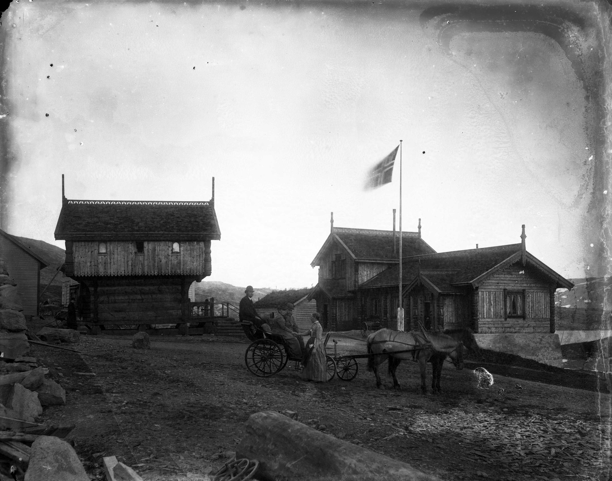 Marthinius Skøien, Gårdstun på Haukeliseter i Telemark, med personer sittende i en vogn trukket av to hester. Bygning, stabbur og flagg i bakgrunnen.
