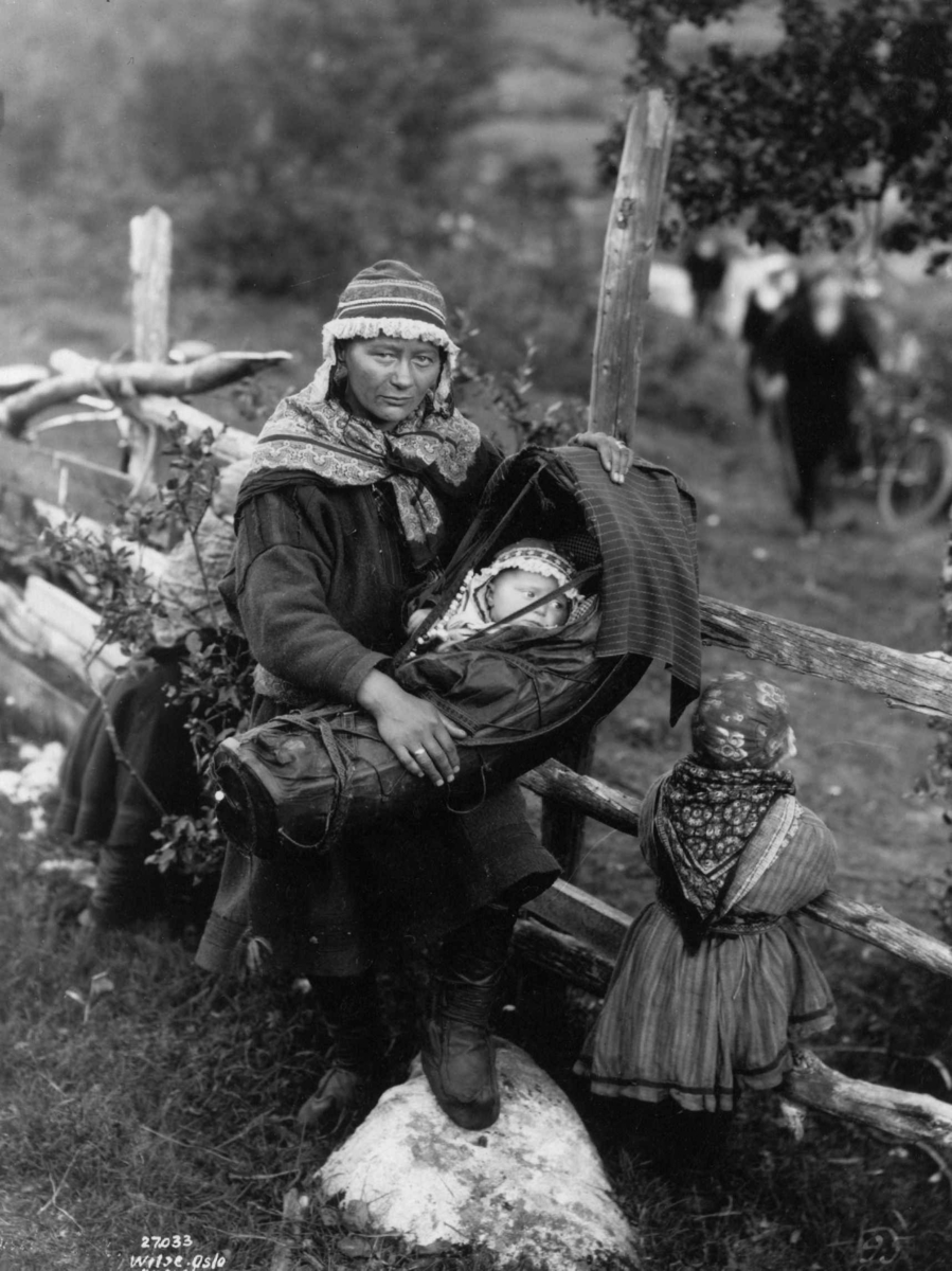 Anders Beer Wilse, En kvinne holder et barn i komse, En liten jente står ved siden av og holder i gjerdet. Antagelig Lyngen 1925