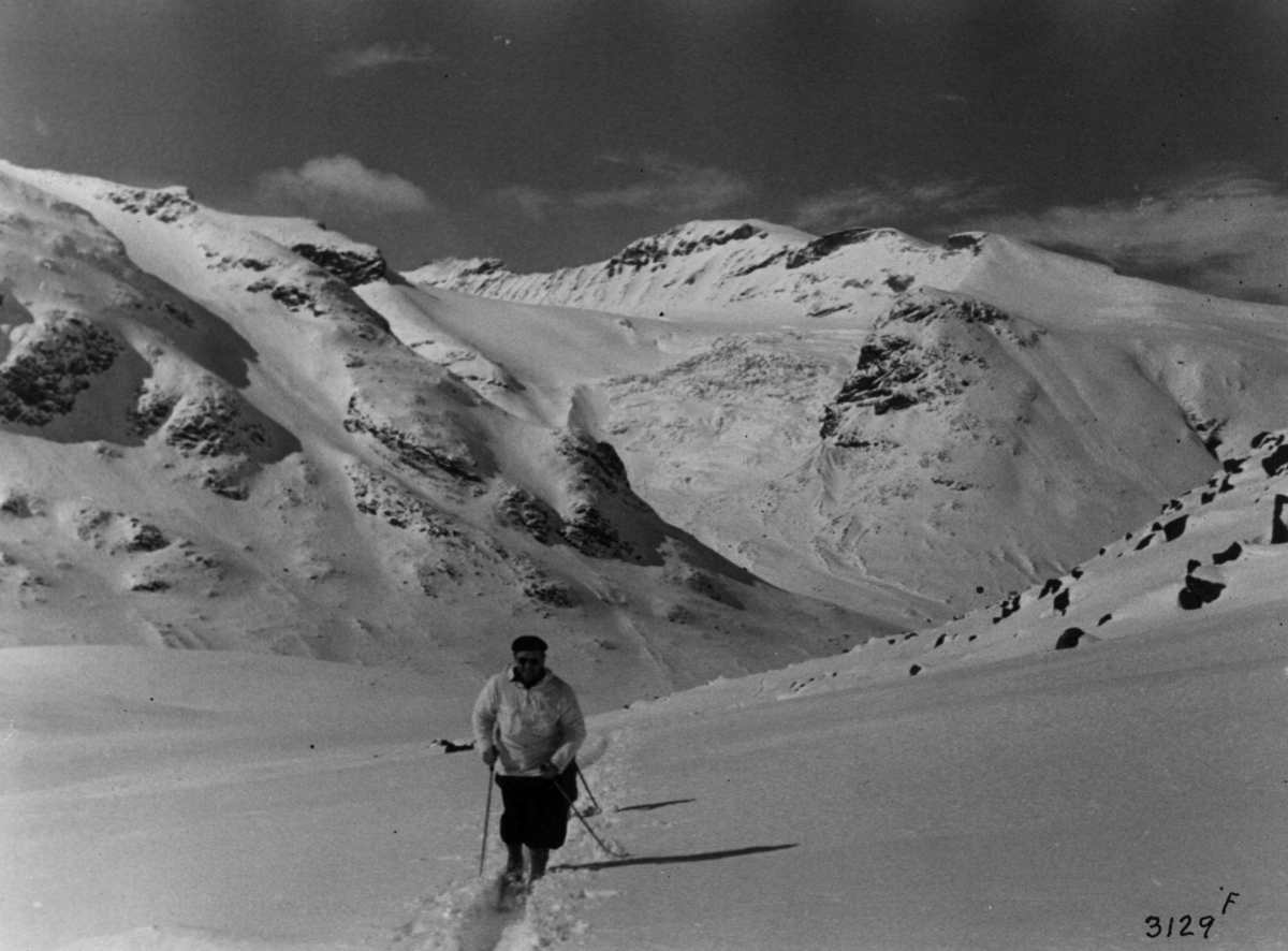 Anders Beer Wilse, Skiløpere ved Galdhøpiggen i Jotunheimen med snødekte fjell og sol.