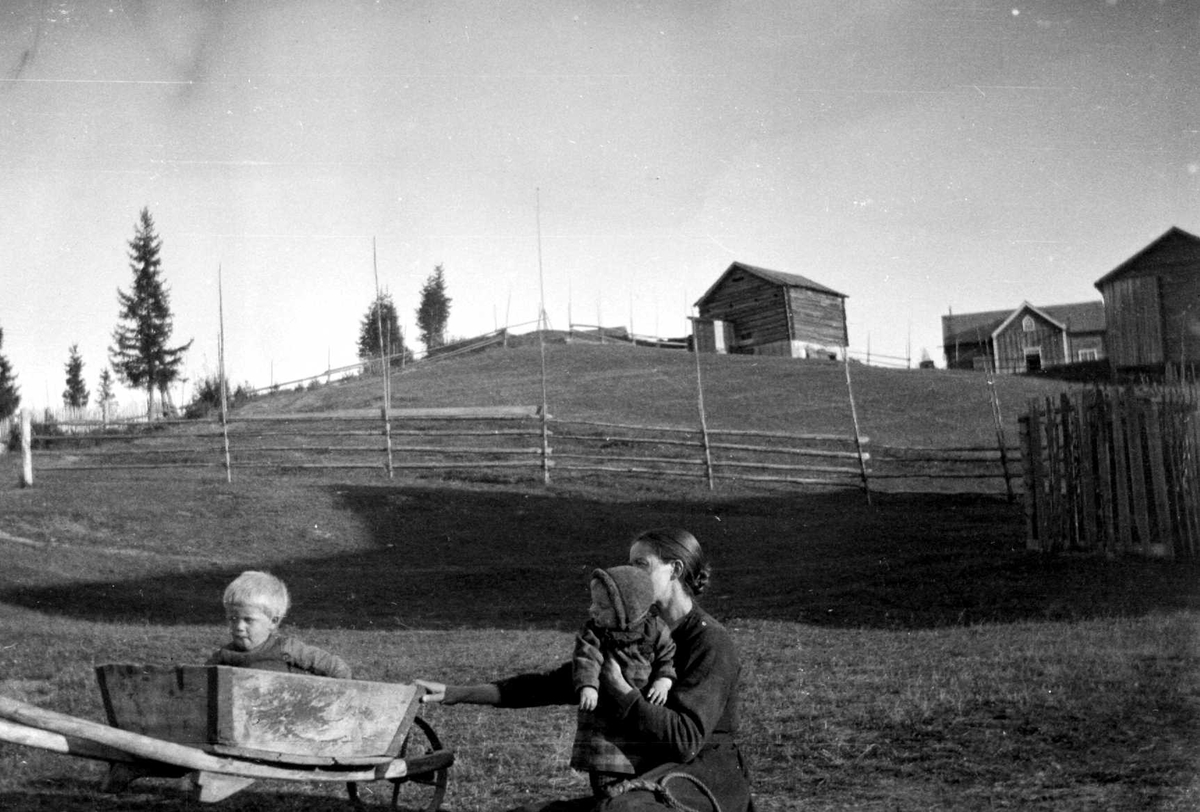 Juliane Fredrikke Langberg, Fra Eggedal, Sigdal hos Th. Kittelsen. Kvinne med to barn, gutten sitter i et trillebør. I bakgrunnen tomme hesjer og gårdsbebyggelse. Fra malerinne Juliane Fredrikke Langbergs (1856-1930) samling tatt ca. 1895-1910 med motivkrets slekt og venner og steder tilknyttet samvær med disse.