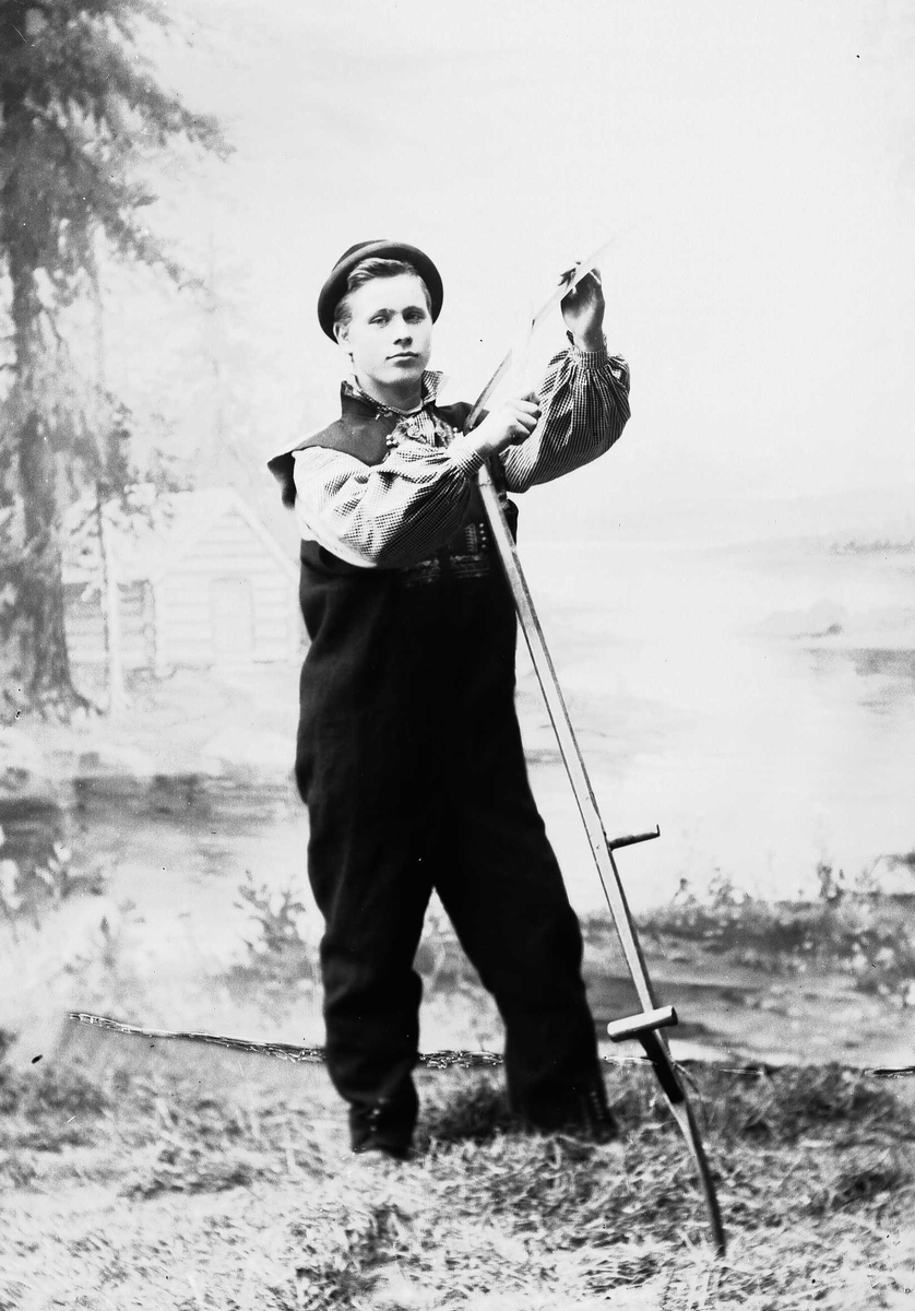 Solveig Lund, Studioportrett av mann i nasjonaldrakt som holder ljå i hendene.Bakgrunnen er malt med skog, vann og hytte. På gulvet er det høy/gress.