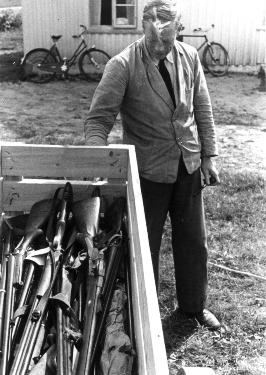 Anna Grostøl, Knut vatnestrøm med en samling våpen i en kasse. To sykler lent mot en husvegg i bakgrunnen. Frostemyr 1941.