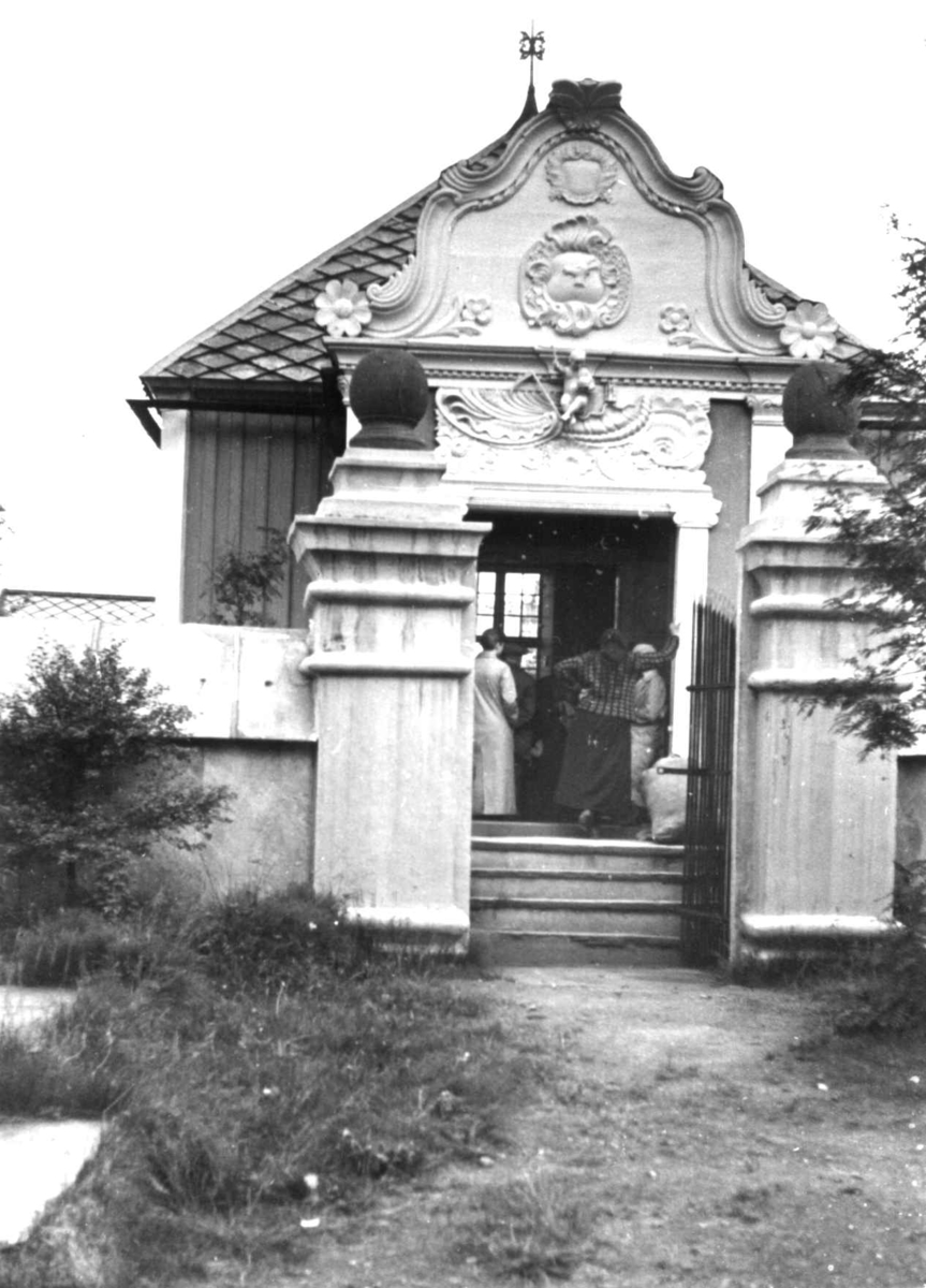 Anna Grostøl, Folk samlet for utdeling av ull i et lite hus med rik ornamentikk, Hiorts gravmæle. Røros 16. juli 1938.