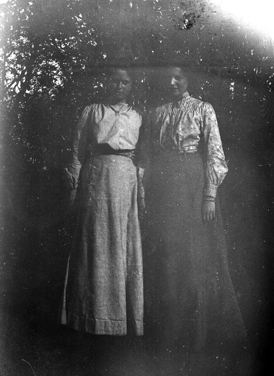 Juliane Fredrikke Langberg, Portrett av 2 stående kvinner, t.v. Emily Holm, fotografens niese. Begge har på seg langt skjørt og bluse. De står foran trær ute, muligens tatt i Hurum eller Rakkestad, hvor Emilys far var prest.. Fra malerinne Juliane Fredrikke Langbergs (1856-1930) samling, tatt ca.1895-1910 med motivkrets slekt…