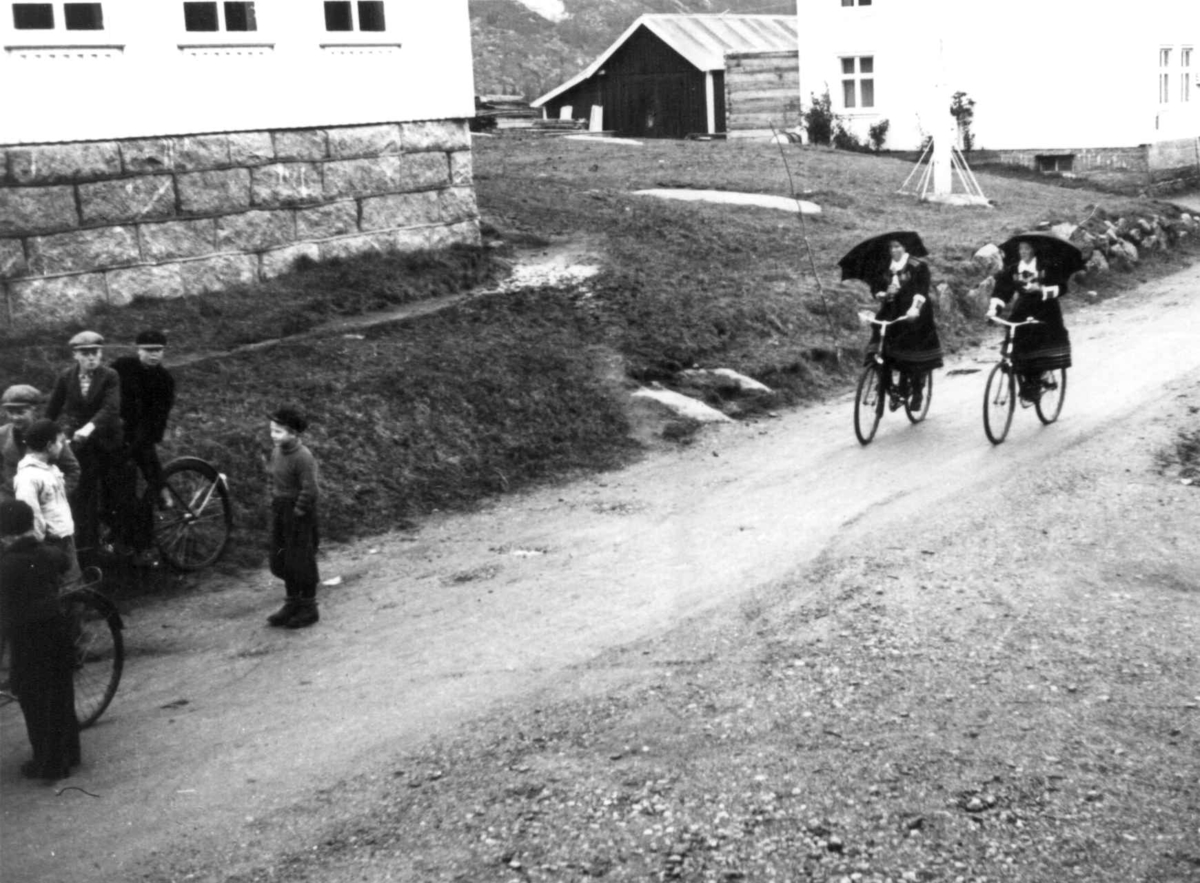Anna Grostøl, Valle, Aust-Agder 1938. Bededag. folk med sykler og parapyer på vei til kirken en regværsdag. Bygninger langs veien.