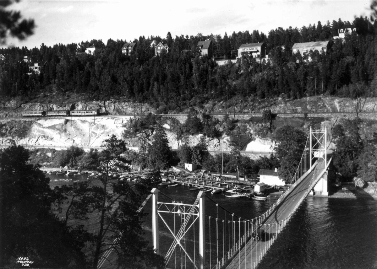 Hermann Christian Neupert, Bekkelaget, Oslo 1937. Brua over til Ulvøya. Bil på brua. Brygger med fortøyde båter ved fjorden nedenfor. Vei og jernbanespor i bakgrunnen opp mot bebyggelse og skog. Tog.