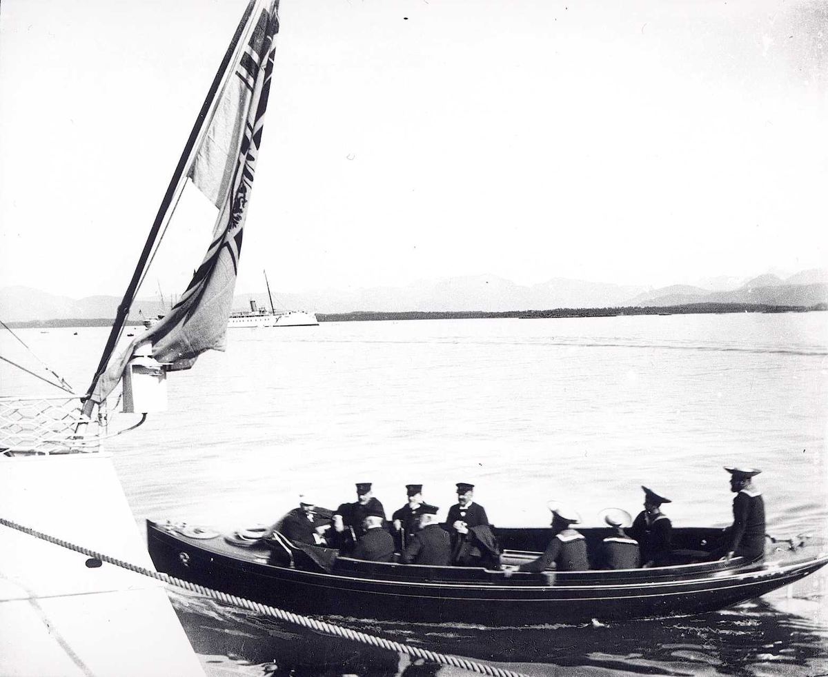 Tom Kent, Motorbåt med uniformrte menn på havna i Molde, Møre og Romsdal, akterstavnen av skip med flagg. Dampskip lenger ut.