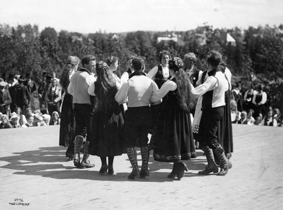 Hermann Christian Neupert, Folkedansoppvisning på en scene. 1925. Publikum. Bebyggelse i bakgrunnen.