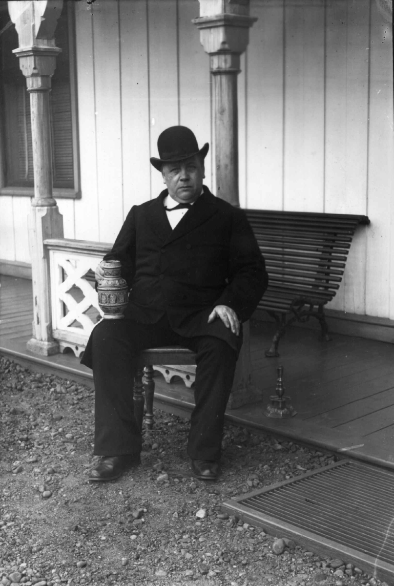 Fredrik Emil Faye, Portrett, lensmann Hansen, Ullensaker, sittende på benk med kanne i keramikk i hånden. Fra portrettserie av personer som bodde på eller besøkte Dal gård, Ullensaker, fotografert av gårdens eier, kammerherre Fredrik Emil Faye (1844-1903) i årene 1875-1900.