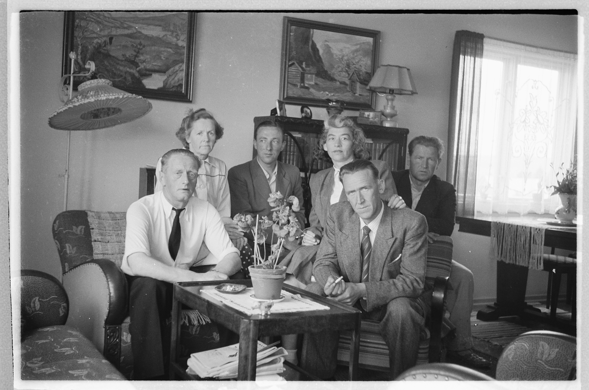 Håkon Prestkværn, Interiør, stue. Ukjente kvinner og menn rundt stuebordet. T. h bak Håkon Prestkværn. Martha (født Prestkværn) og Paul Pettersen, begge i lyse klær. Bildet er tatt i Martha og Pauls stue i leiligheten i Lillestrøm. Håkon sitter bak på motsatt side.