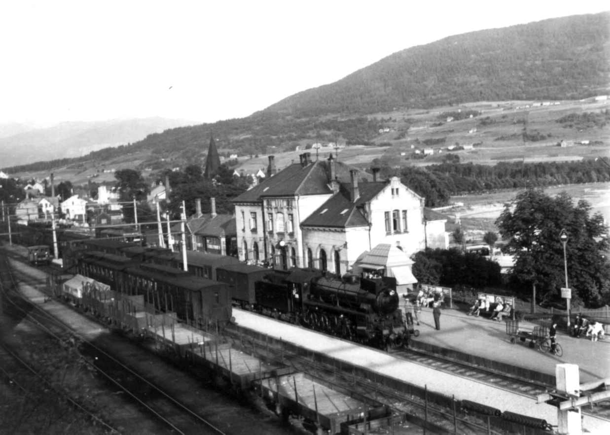 Hermann Christian Neupert, Voss jernbanestasjon 1939. Tog med damplokomotiv på stasjonen. Godsvogner. Tilskuere. Bebyggelse. Gårder i bakgrunnen. Skog og fjell.