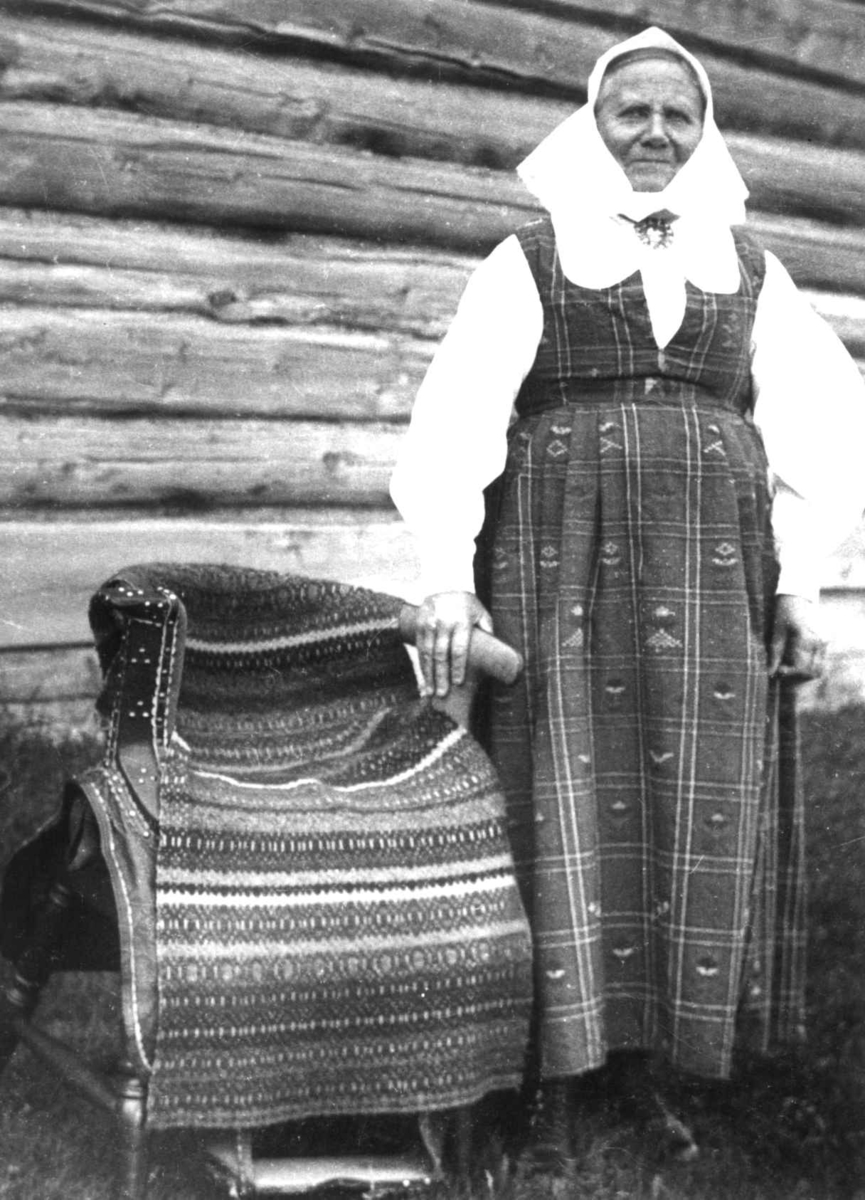 Anna Grostøl, Antagelig Lesja, Oppland 1934. Kvinne kledd i kvinnedrakt, huns står ved siden av en stol med teppe.