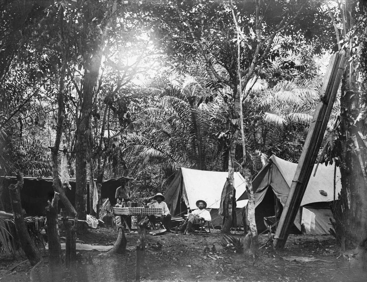 Gustav Borgen, Avfotografering. Teltleir i jungelen. Tre menn rundt et dekket bord.