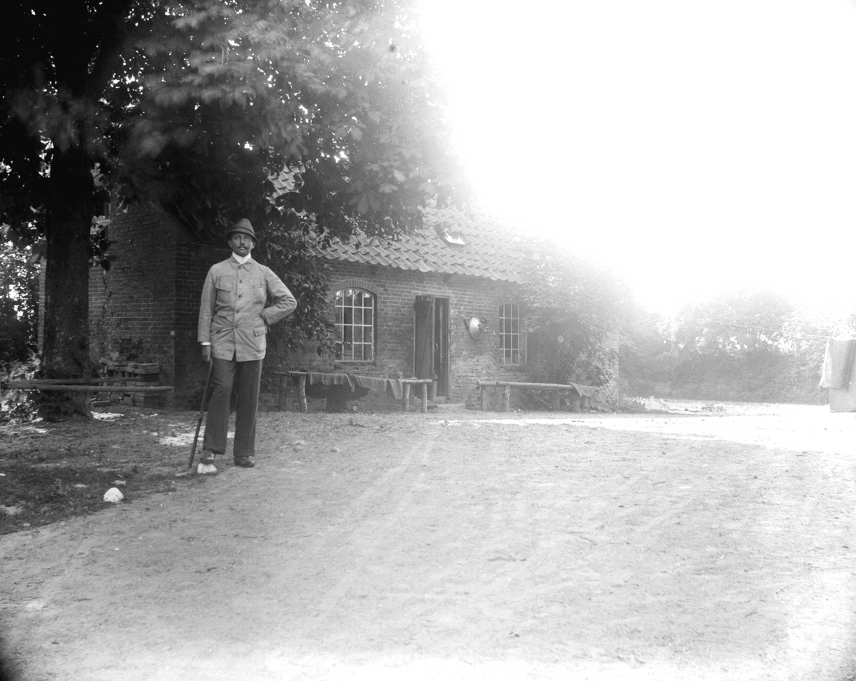 Juliane Fredrikke Langberg, Portrett av Heinrich C Hudtwalcker, g. 1903 med Sigrid f. Holm, fotografens niese. Han står med stokk foran lav murbygning, ant. i Hamburg. Fra malerinne Juliane Fredrikke Langbergs (1856-1930) samling, tatt ca.1895-1910 med motiver fra slekt og venner samt steder tilknyttet samvær med disse.