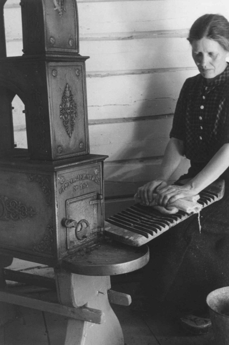 Anna Grostøl, en kvinne tover på et tovebrett ved siden av en etasjeovn til vedfyring. Åseral, Vest-Agder 1943.