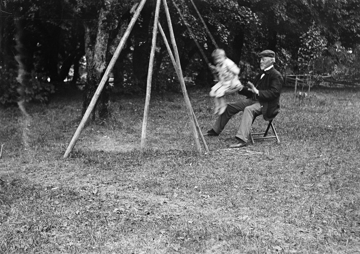Okänd,, Ruben och Christiane Liljefors son Roland med sin morfar Roland Petersen, gungar ute i trädgården, sannolikt i Kärrbogärde 1906