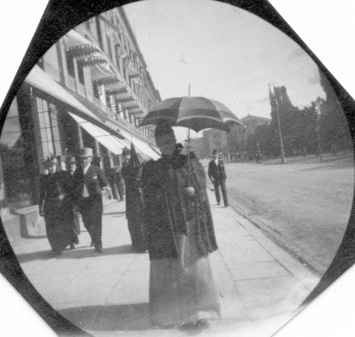 Fredrik Carl Mülertz Størmer, Fru apoteker Størmer, fotografens mor, spaserer langs Karl Johans gate med parasoll. Stortinget skimtes.