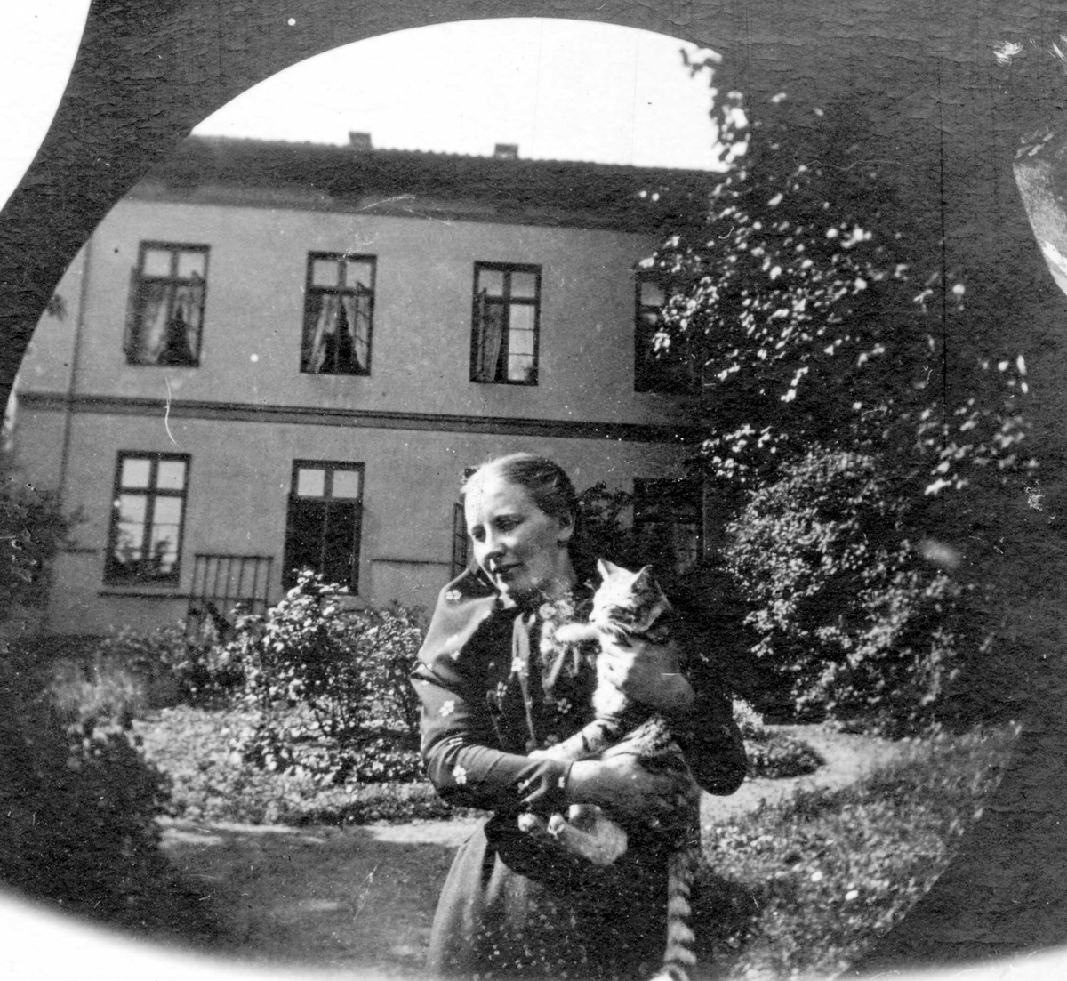 Fredrik Carl Mülertz Størmer, Professor Blytts stuepike og katt. Kvinne med stipet katt på armen i hage forann toetasjers hus, Oslo.
