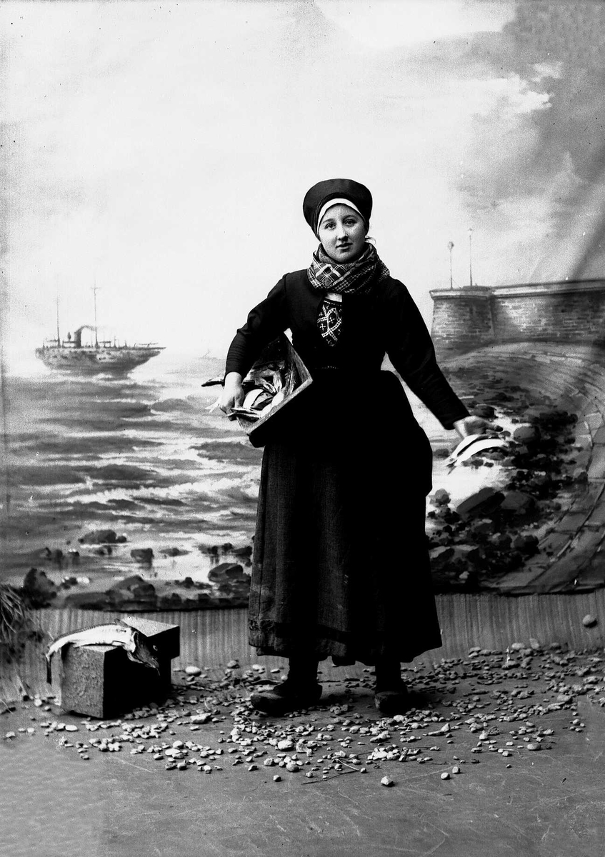 Solveig Lund, Studioportrett av kvinne i drakt med lue på hodet. Til høyre for henne ligger fisk på blokk. Hun holder et trau med fisk under den ene armen og to fisker i den andre. Bakgrunnen er malt med sjø, himmel, steiner, dampbåt og pir.