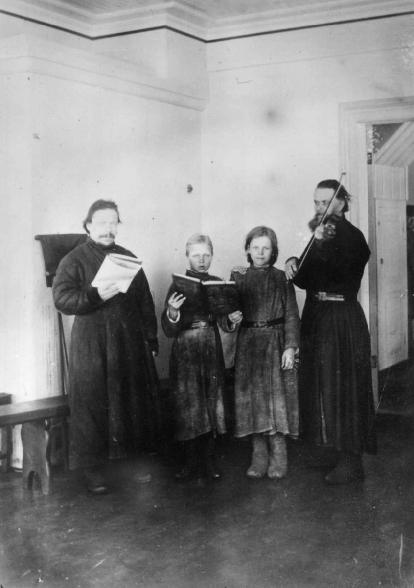 Ellisif Rannveig Wessel, Klosteret i Petsjenga (Petsamo), Russland, interiør med to menn og to piker, mannen t. h. med fiolin, ca. 1900.