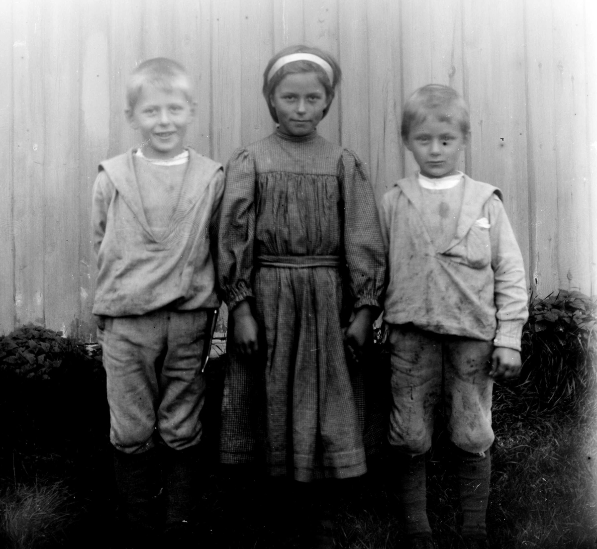 Juliane Fredrikke Langberg, Portrett av 3 barn, fra v. Olaf, Emily og Kristofer Holm, barn av res. kap. i Rakkestad 1884-97, Olaf A. Holm, gift med fotografens søster Louise. Barna står foran en husvegg. Ant. fra kapellangården Bergenhus i Rakkestad. Fra malerinne Juliane Fredrikke Langbergs (1856-1930) samling, tatt ca.1895-…