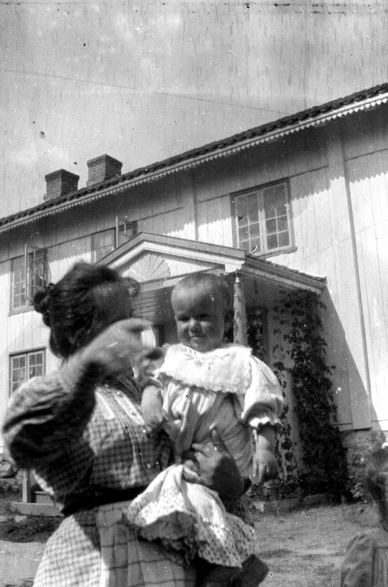 Juliane Fredrikke Langberg, Fra gårdstun i Hurum foran våningshus i 2 etasjer med inngangsparti midt på (som NF.15386-036). Kvinne med et barn på armen i forgrunnen (ukjente). Fra malerinne Juliane Fredrikke Langbergs (1856-1930) samling, tatt ca.1895-1910 med motivkrets slekt og venner samt steder tilknyttet samvær med disse.