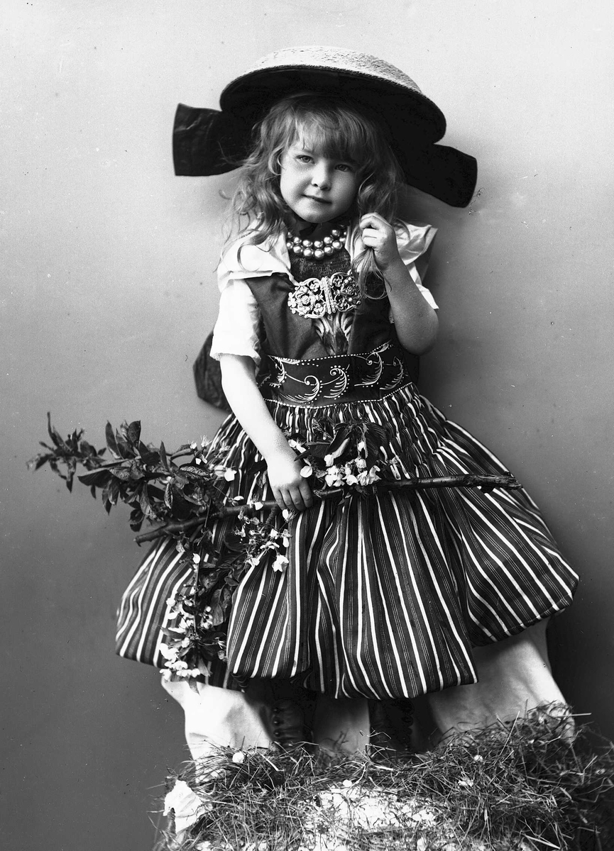Solveig Lund, Pike i drakt og hatt, med perlekjede i halsen. Holder gren med blader og blomster.