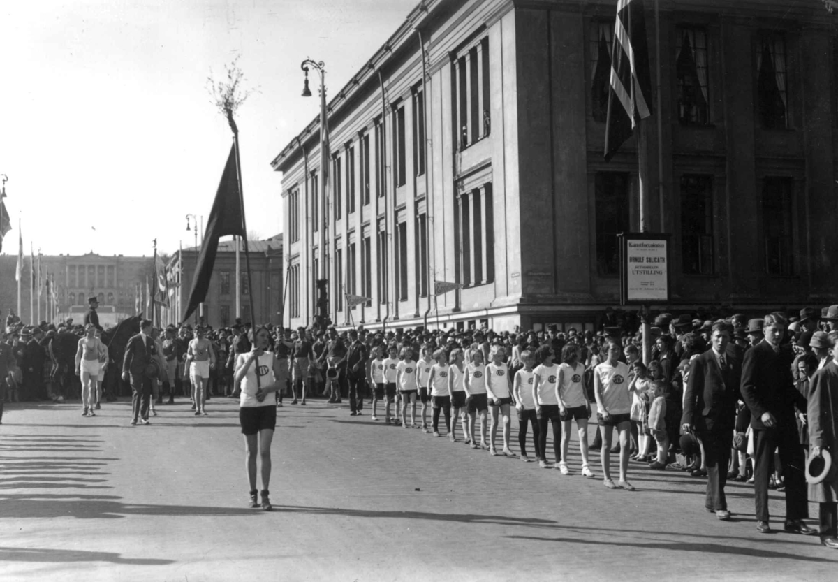 Hermann Christian Neupert, 1. mai-tog, Karl Johans gate ved Universitetet. 1930. Mann med fane. Barn og menn i sportsantrekk. Slottet i bakgrunnen. Tlskuere.