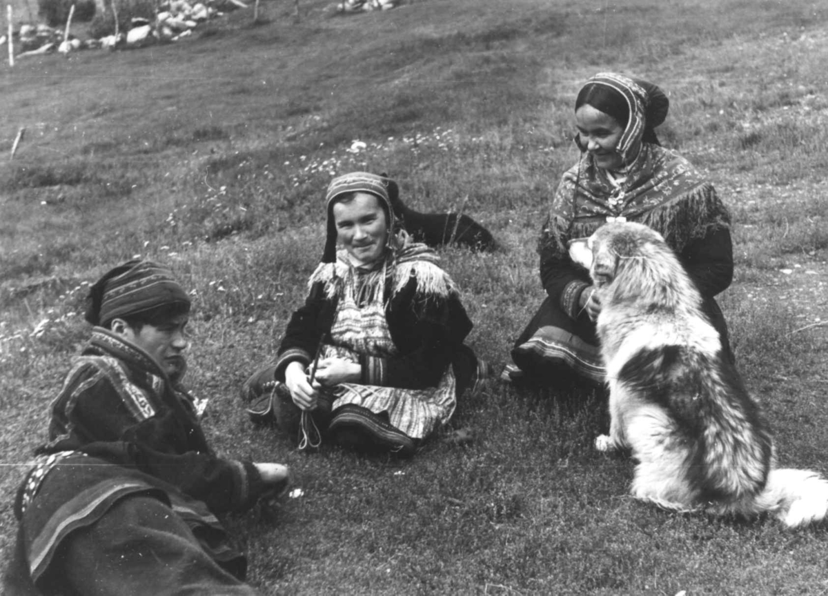 Anna Grostøl, Samer sitter i gresset sammen med en hund. Olderdalen, Kåfjord, Troms 1947.