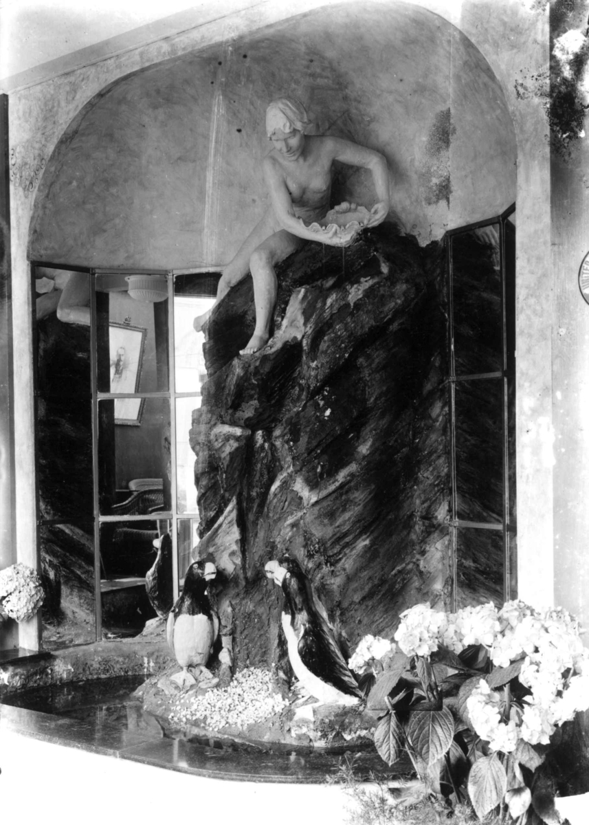 Hermann Christian Neupert, Jubileumsutstillingen på Frogner, Oslo, 1914. Interiør paviljong. Skulptur av kvinne. To lundefugler foran denne. Blomster i forgrunnen.
