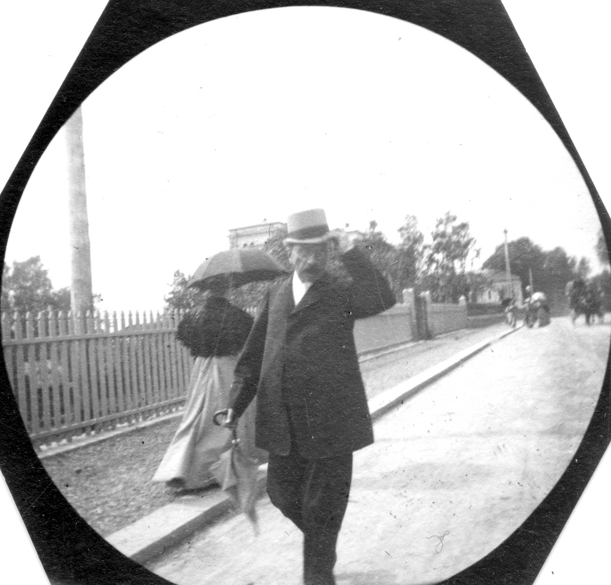 Fredrik Carl Mülertz Størmer, Overlærer Wallem, med hånden på hatten, spaserer i gate langs gjerde.Hestevogn og villa i bakgrunnen.