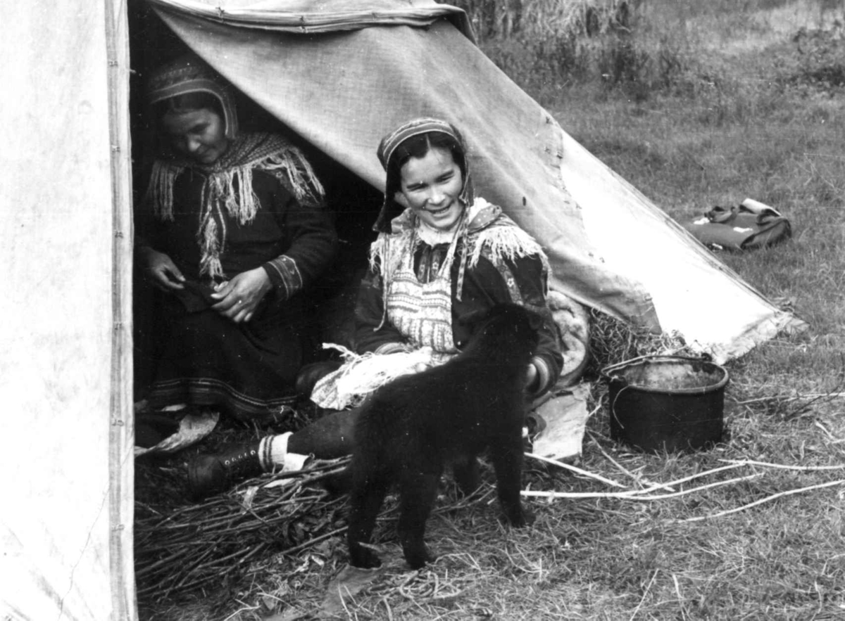 Anna Grostøl, To kvinner i teltåpningen arbeider med skinn og senetråd, en hundehvalp i forgrunnen. Olderdalen, Kåfjord, Troms 1947.