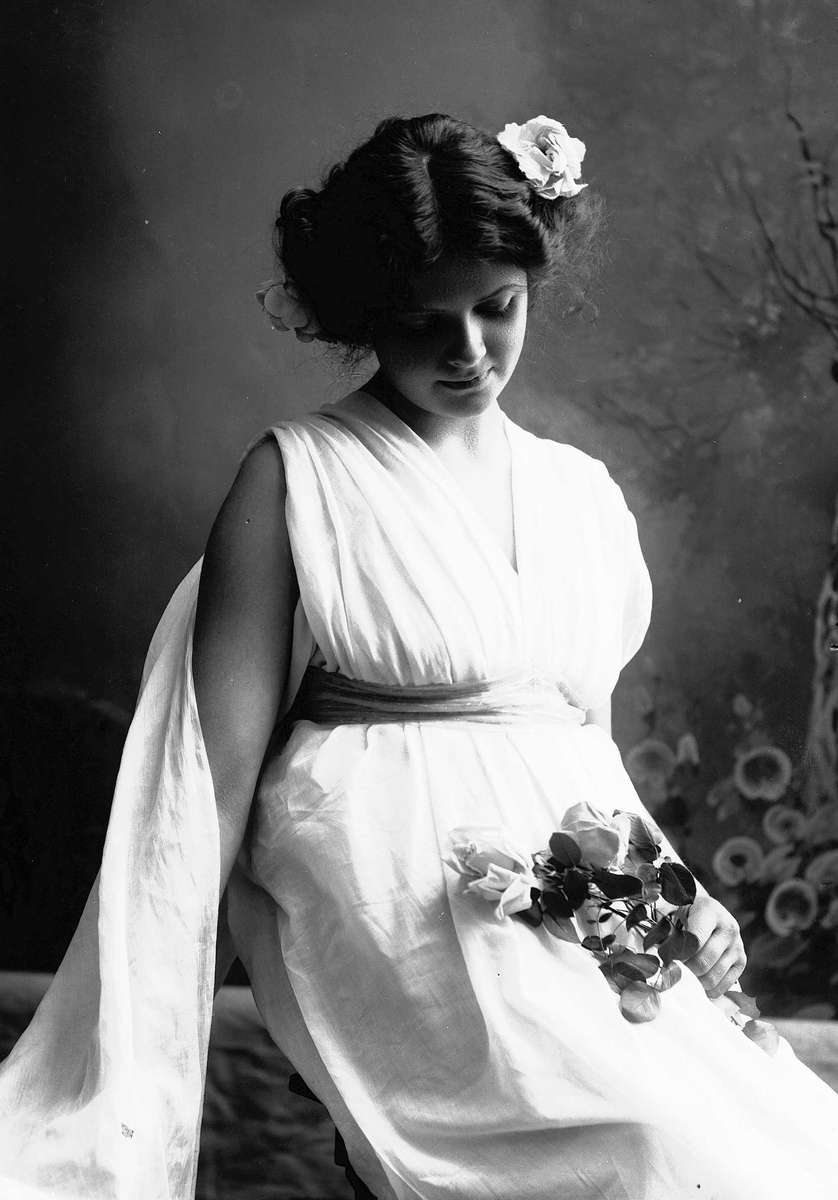 Solveig Lund, Studioportrett av kvinne i kostyme med hvit kjortel og blomster i håret. I den ene hånden holder hun en blomsterbukett og bakgrunnen er malt med trær og blomster.