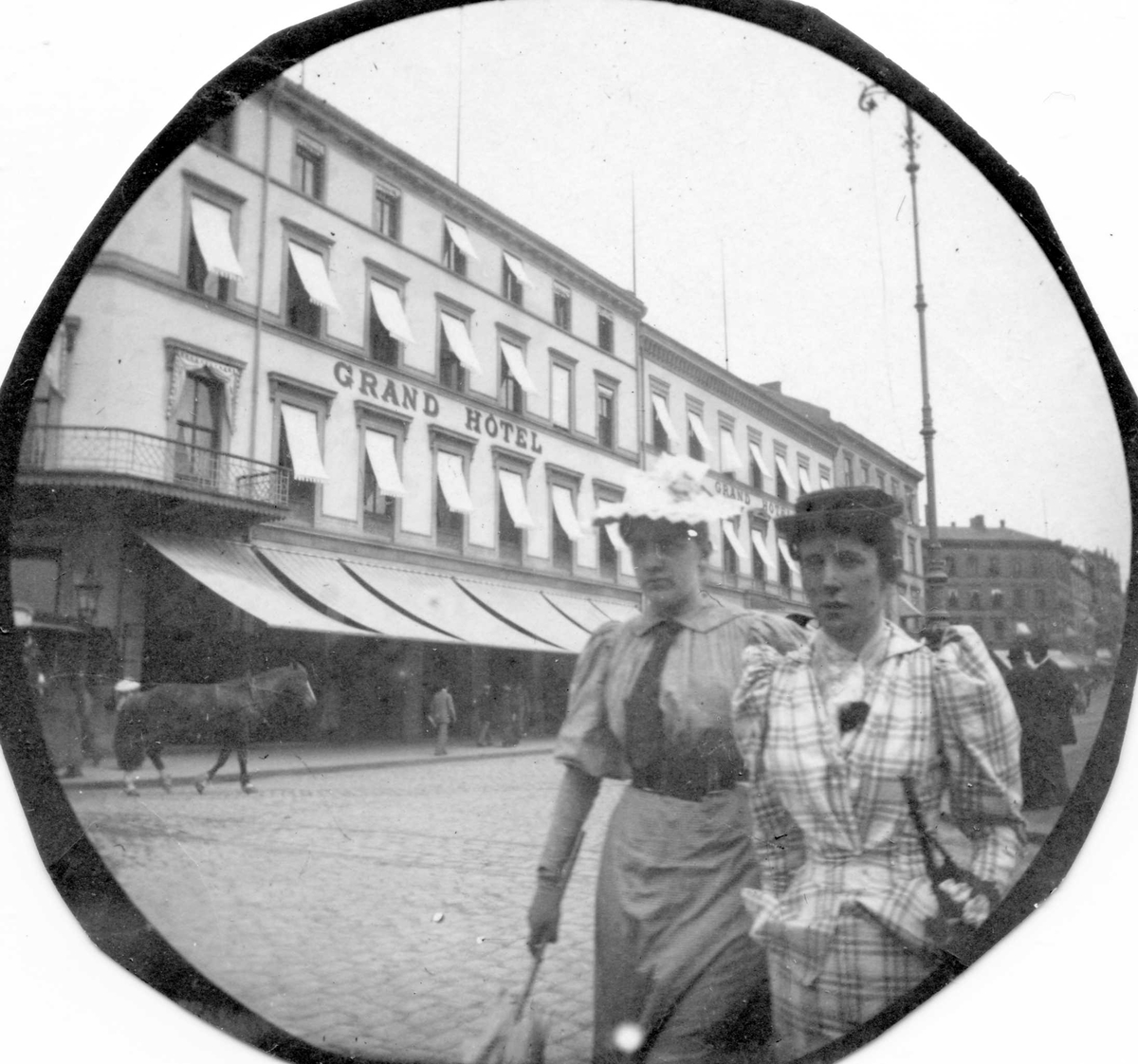 Fredrik Carl Mülertz Størmer, Frk. Just senior og en annen kvinne spaserer langs Karl Johans gate, Oslo, på andre siden for Grand Hotell.