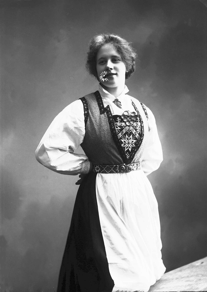 Solveig Lund, 3/4 del studioportrett av kvinne i drakt med oppsatt hår. I munnen har hun en prestekrage og hun holder hendene på hoftene. Det høyre benet hviler mot en kuliss. Kvinnen ser ut av fotografiet. Nøytral bakgrunn.