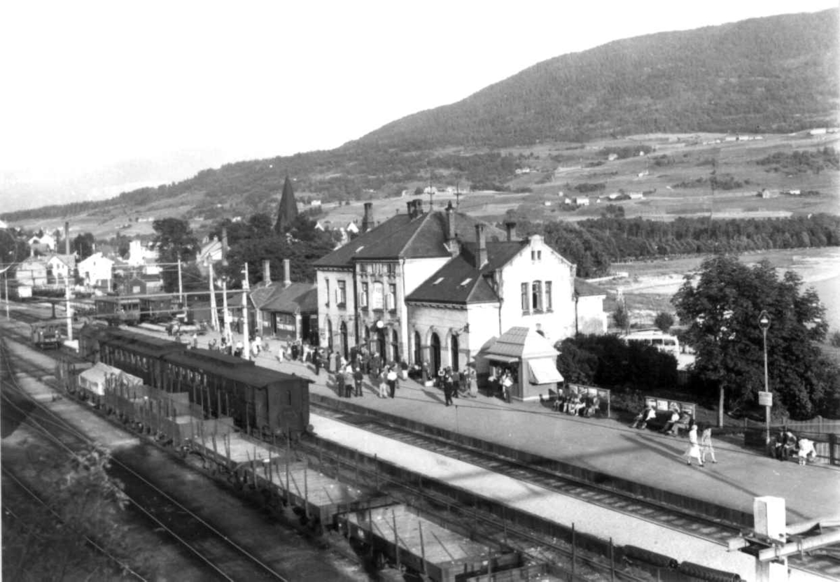 Hermann Christian Neupert, Voss jernbanestasjon. 1939. Jernbanevogner og godsvogner på stasjonen. Passasjerer på perrongen. Landskap med gårder, jorder og åkrer. Skog og fjell i bakgrunnen.
