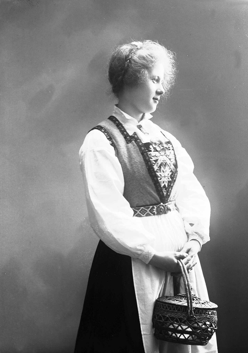 Solveig Lund, 3/4 dels studioportrett av kvinne i drakt. I hendene holder hun en stråkurv.