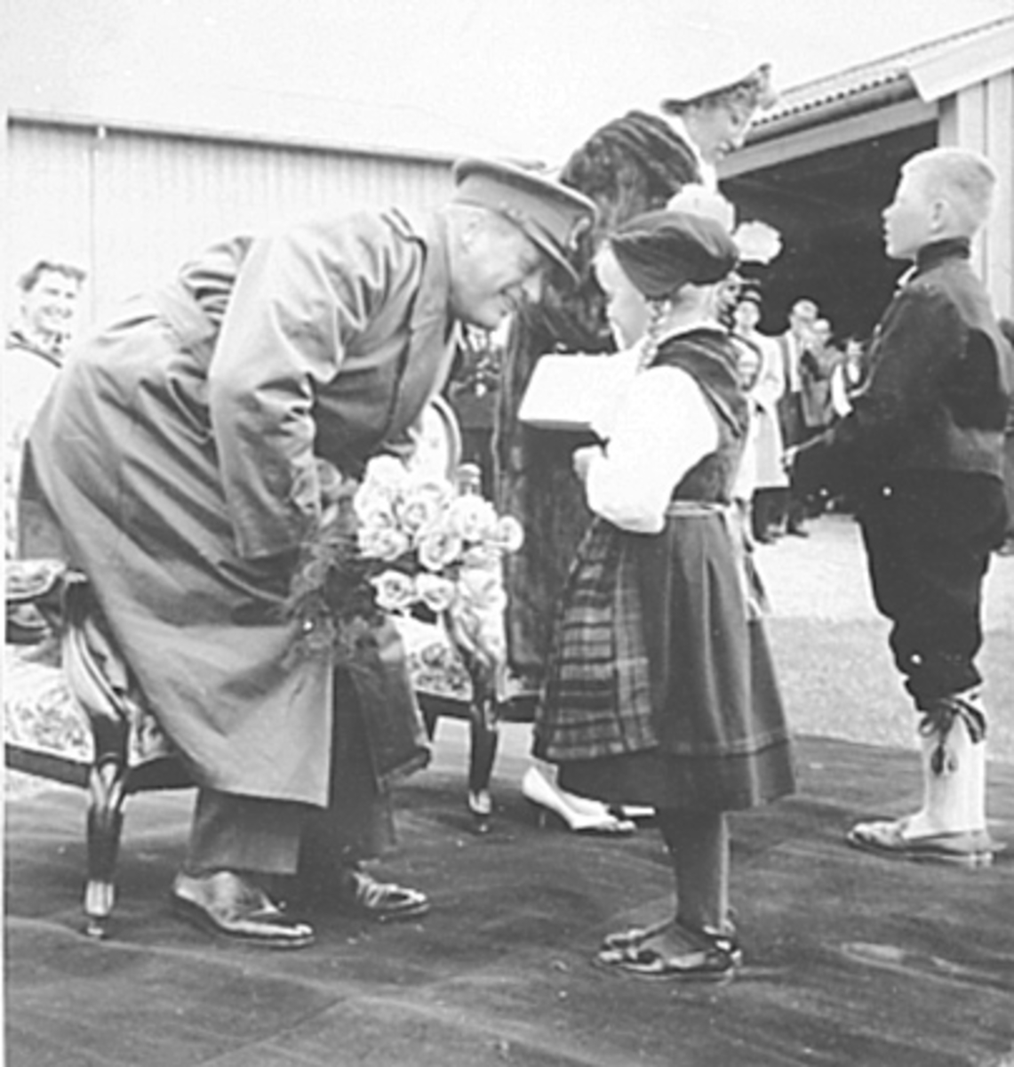 Anno Domkirkeodden, HKH KONG OLAV V OG PRINSESSE ASTRID, SIGNINGSFERDEN, MOTTAR BLOMSTER KONG OLAV TAR IMOT BLOMSTER AV EN PIKE I BUNAD. PRINSESSE ASTRID I BAKGRUNNENEN I SAMTALE MED EN LITEN GUTT