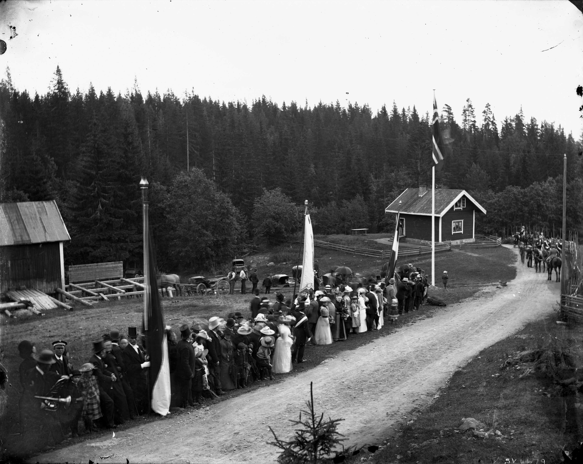 Marthinius Skøien, Oppstilt, festkledd folkemengde ved vei med flagg og faner, ukjent sted og anledning. Ridende soldater i opptog til høyre?