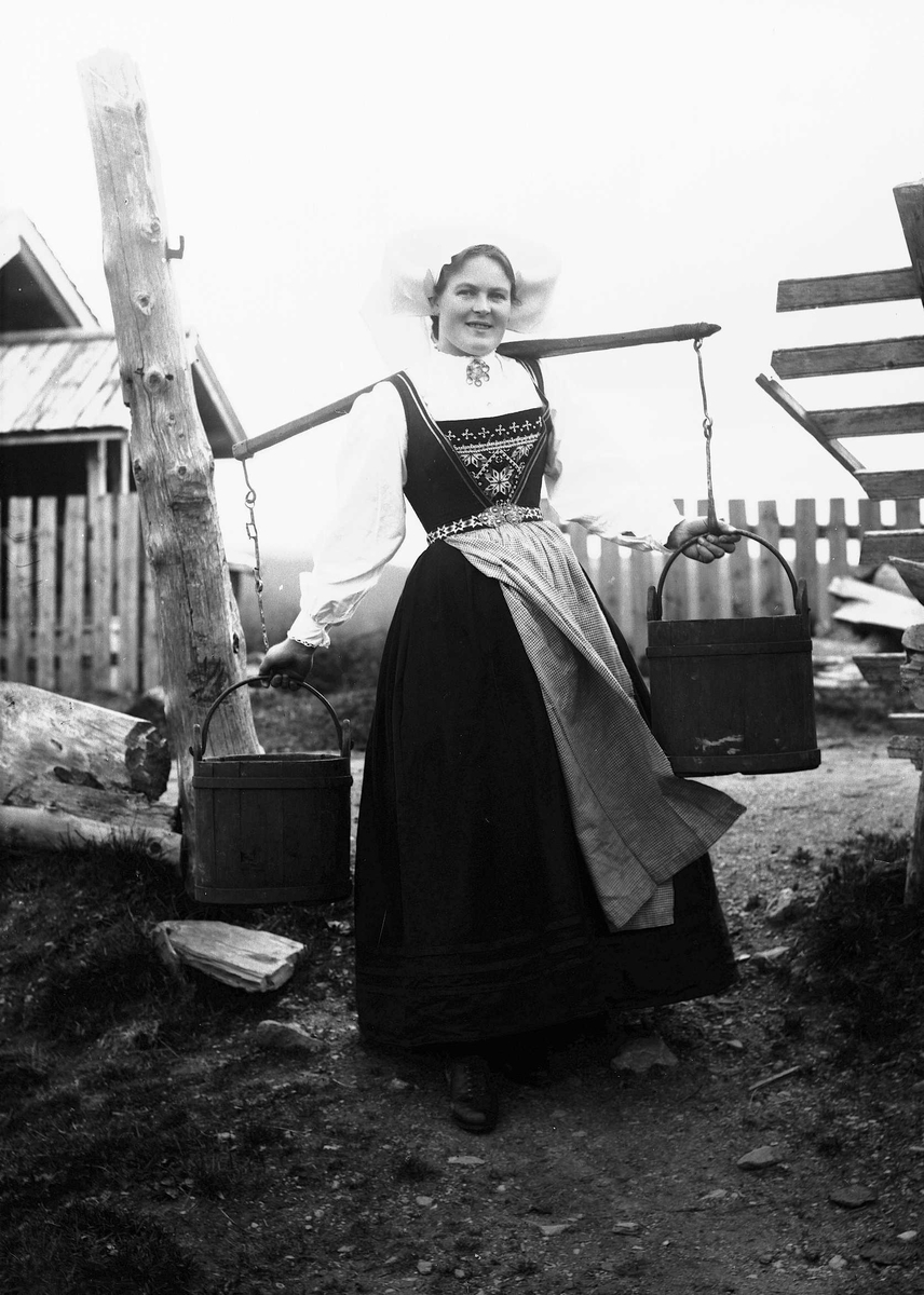 Solveig Lund, Kvinne i Hardangerdrakt står utenfor seter. Bærer på to vannbøtter.