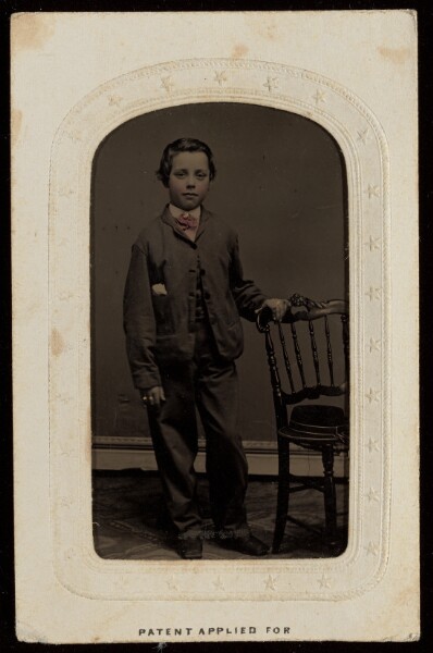 Unknown, [Unidentified little boy wearing a tie standing, one hand to a chair back]