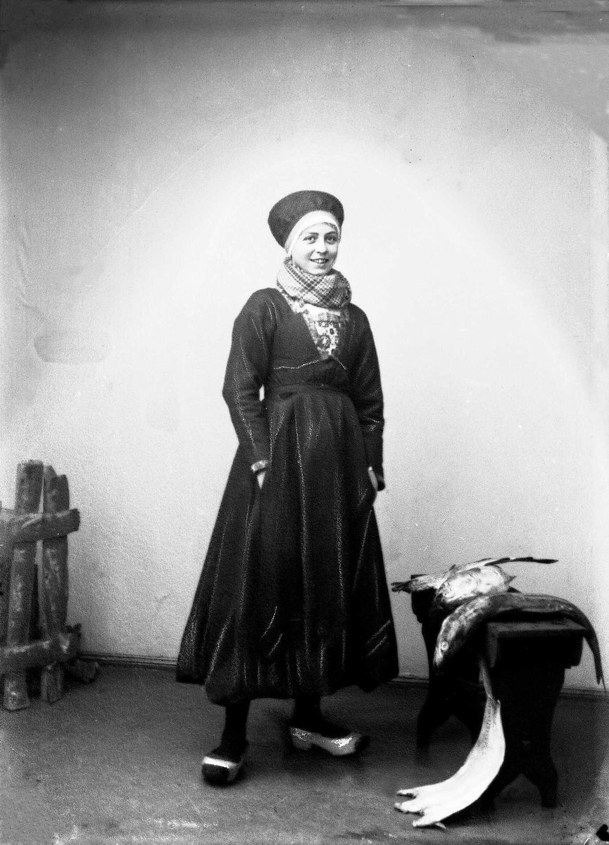 Solveig Lund, Studioportrett av kvinne i drakt, skaut/hatt, tresko og sjal i halsen. Til høyre for henne står en benk der det ligger flere fisker.Til venstre står et gjerde.