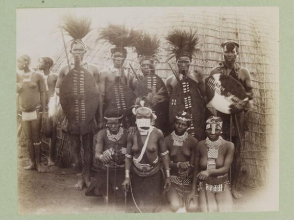 Unknown, A group portrait of (?) Zulu men and women in tribal dress. They are posed in front of a hut.