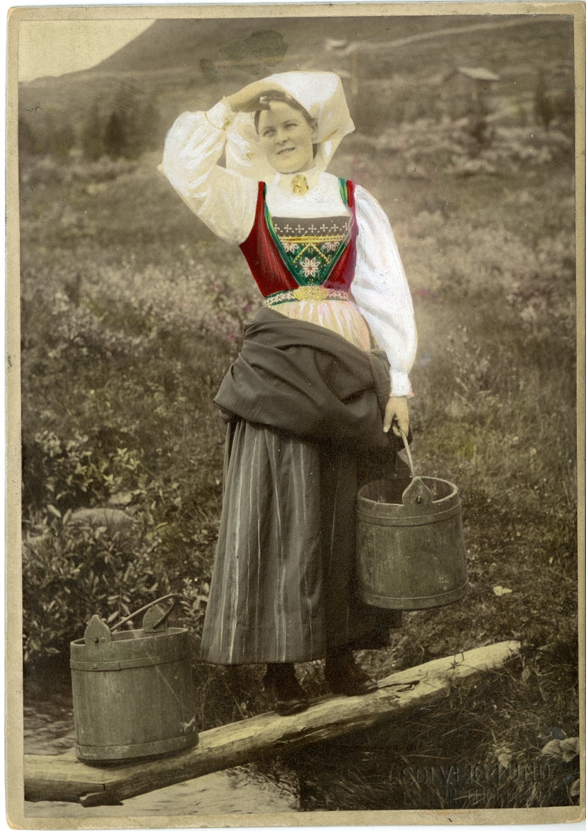 Solveig Lund, Kolorert fotografi av kvinne med drakt, på stokk over bekk bærende på to bøtter. Hardanger.