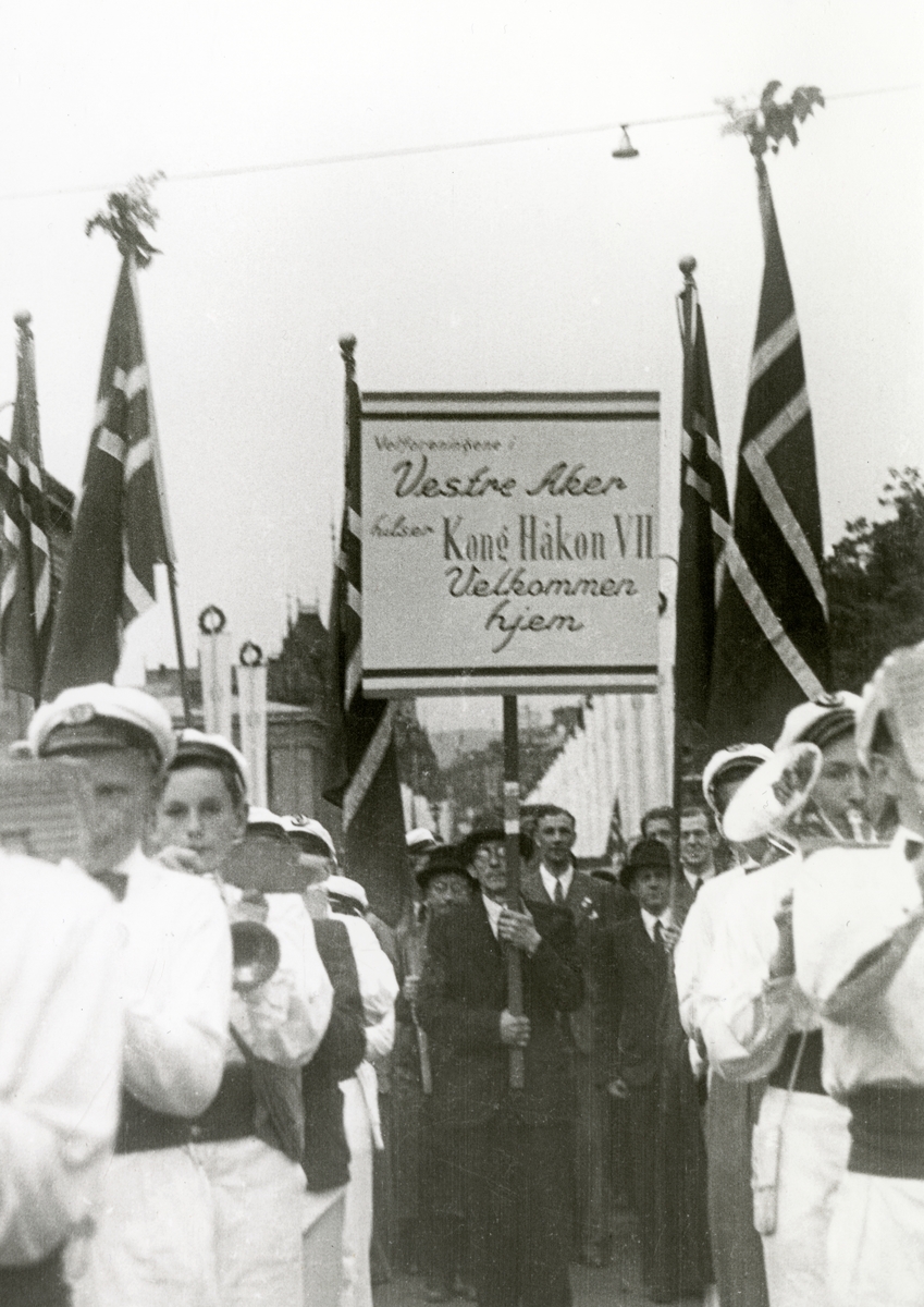 Anders Beer Wilse, Fra Oslo under fredsdagene i 1945. Velforeningene i Vestre Aker, marsjerer oppover Karl Johans gate, med plakat som ønsker kongen velkommen hjem. Musikkorps i forgrunnen.