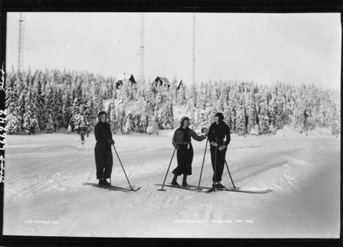 Hermann Christian Neupert, Tryvann, Oslo. Vintermotiv.Tre skiløpere i løypa. I bakgrunnen ses Tryvanns-mastene og bebyggelse.