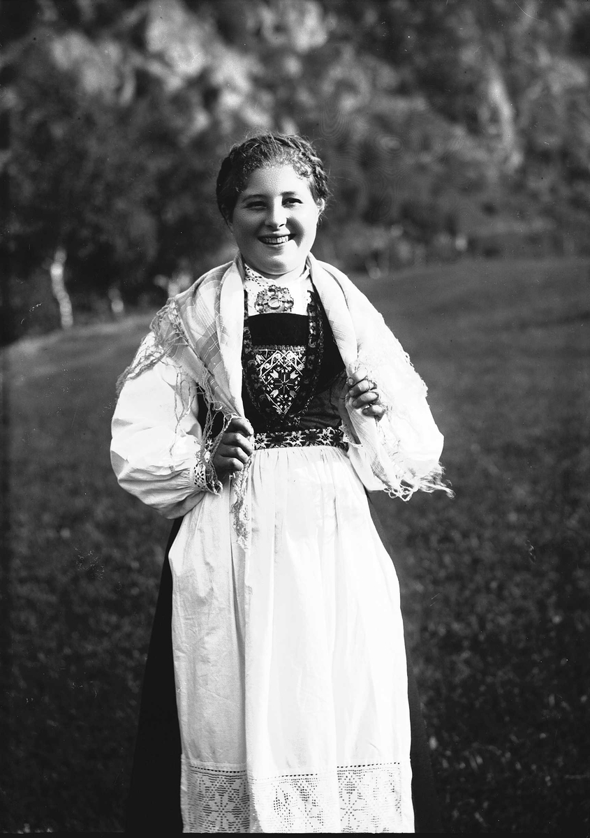 Solveig Lund, Kvinne i nasjonaldrakt står på jorde/eng. Fjell i bakgrunnen. Holder i endene på skautet sitt