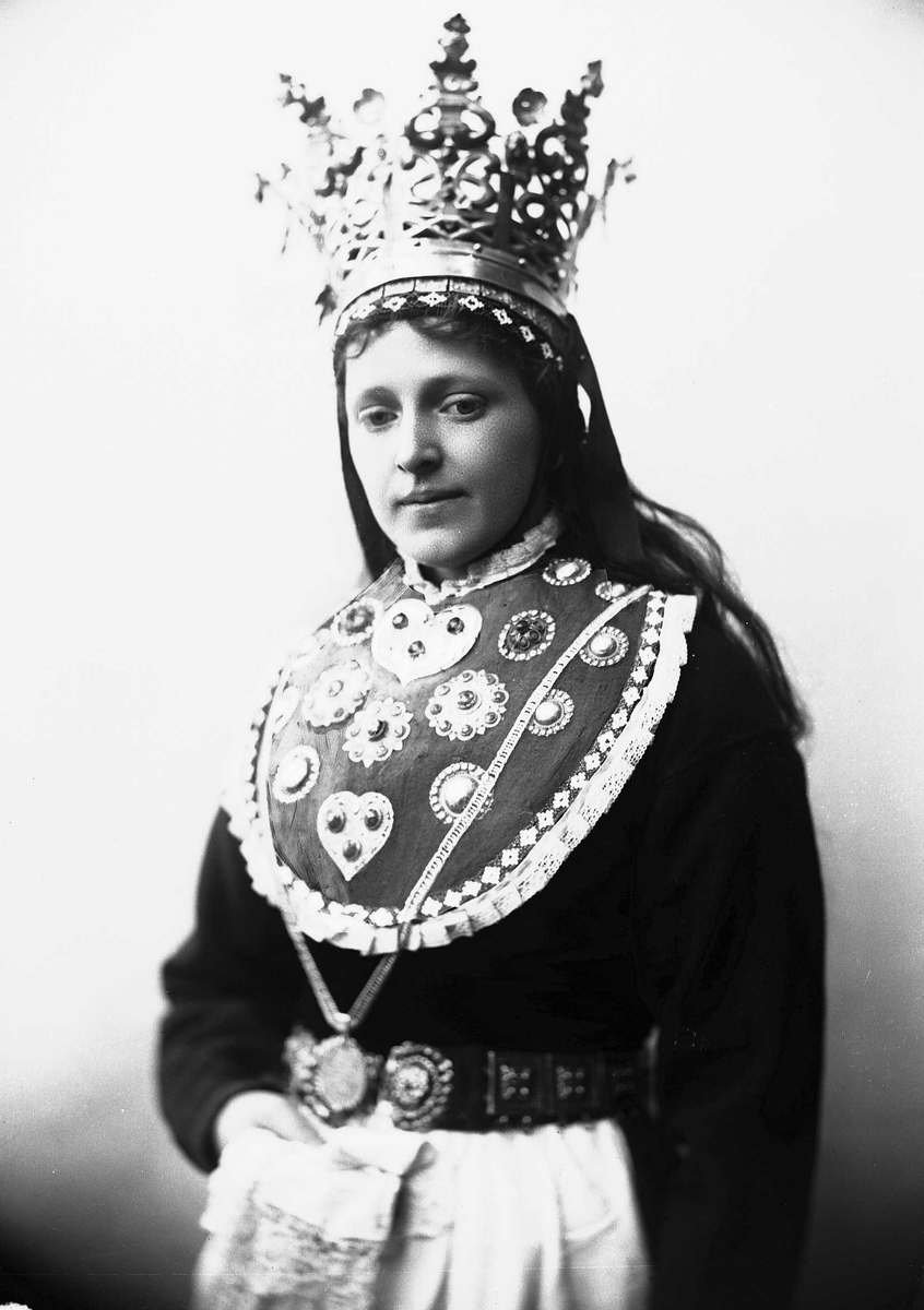 Solveig Lund, Studiofotografi av kvinne med brudedrakt og brudekrone fra Sogn. Holder klede i hånden. Utslått hår. Hofteutsnitt. Vendt mot høyre. Ser nedover.