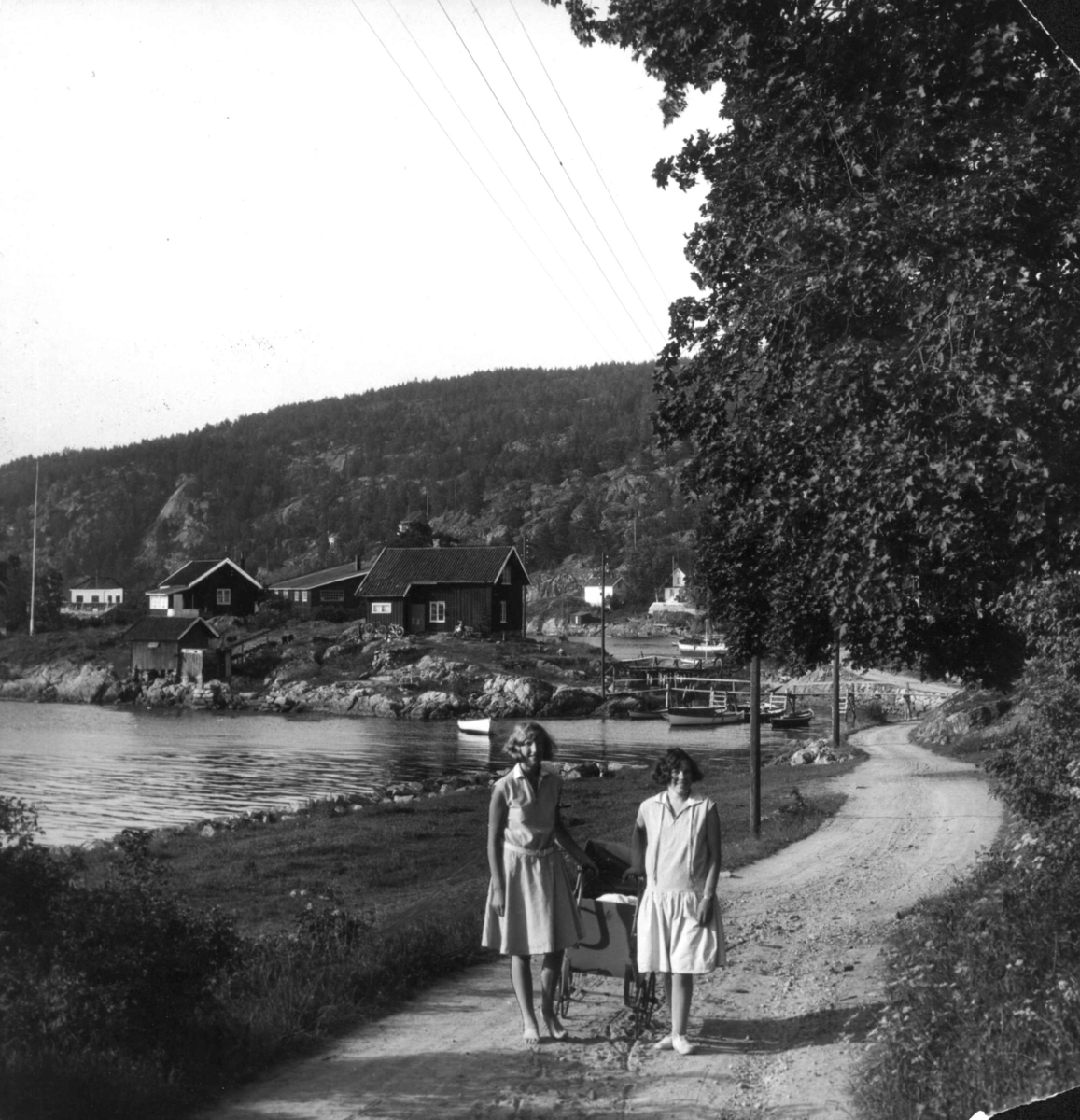 Hermann Christian Neupert, Drøbak, Akershus 1929. To unge kvinner i sommerkjoler med barnevogn står på en vei som går langs sjøen. Bebyggelse og båter ses i bakgrunnen.